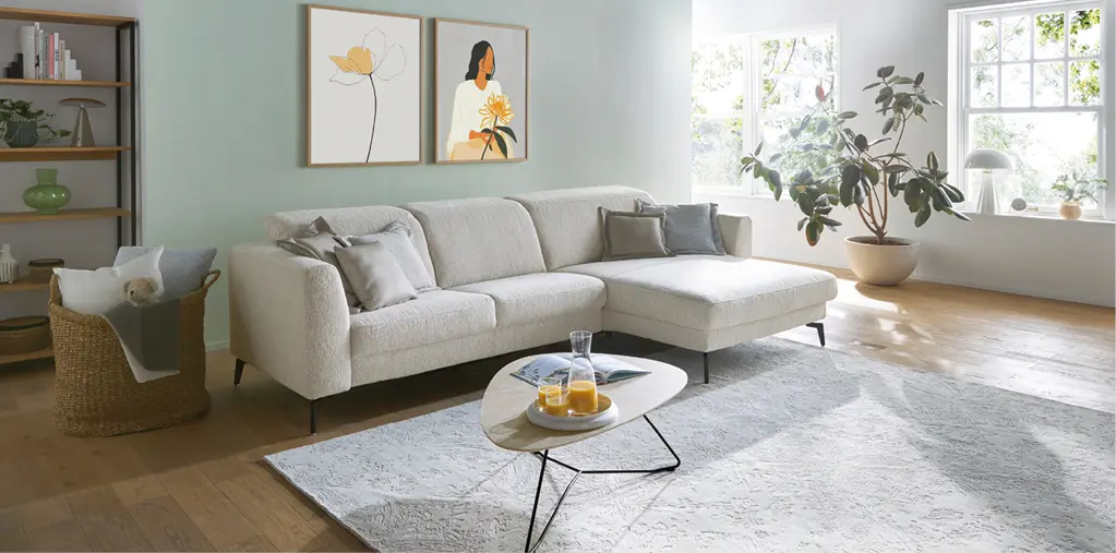 Helles Wohnzimmer mit modernem Ecksofa in Beige, großem Teppich und Couchtisch. Freundliche Einrichtung mit Pflanzen, Tageslicht und ruhigen Farben für ein gemütliches und stilvolles Wohnen.